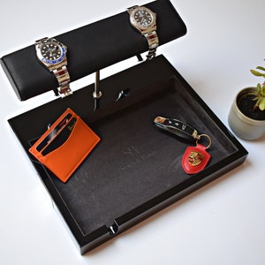 May include: A black valet tray with a gray felt lining. The tray has a watch winder with two silver watches, a leather card holder, a key with a red leather fob, and the text "SR, Small Affluence" on the felt lining.