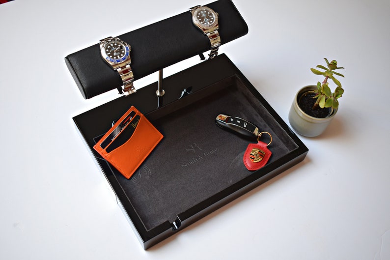 May include: A black valet tray with a gray felt lining. The tray has a watch winder with two silver watches, a leather card holder, a key with a red leather fob, and the text "SR, Small Affluence" on the felt lining.