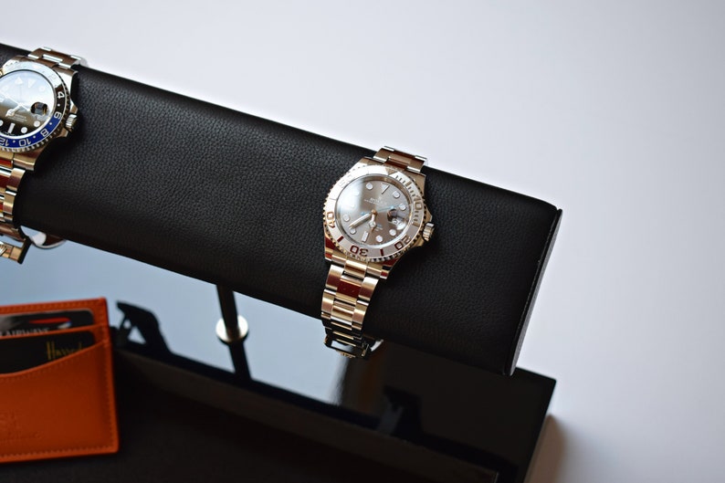 May include: Two silver wristwatches with black leather watch bands on a black display stand. The watch on the left has a blue bezel and the watch on the right has a silver bezel.