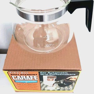May include: A clear glass carafe with a silver rim and black handle, sitting on a brown cardboard box. The carafe has markings for cup measurements. The box reads "FITZ-ALL REPLACEMENT CARAFE" and "For Automatic Drip Coffeemakers".