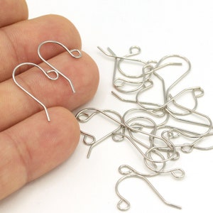 May include: Silver earring hooks, a pile of hooks on a white surface, and two hooks held in a hand.