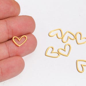 May include: A collection of small, gold-colored heart-shaped charms. One charm is held in a hand, while several others are scattered on a white surface. These delicate charms are ideal for jewelry making or craft projects.