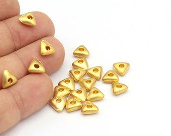 May include: A group of gold triangle-shaped beads with a hole in the center. The beads are scattered on a white surface.
