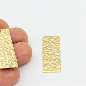 May include: Two gold-colored metal rectangular pendant charms with a hammered texture. The charms have a hole at the top for attaching to a chain or cord.