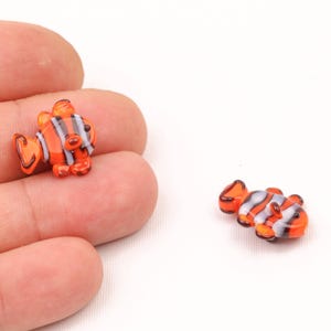 May include: Two small glass figurines of orange and white clownfish. The figurines are detailed with black stripes and have a realistic appearance.