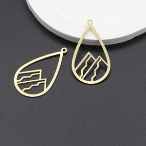 May include: Two gold-colored metal pendant charms in the shape of a teardrop. One charm features a geometric mountain design, while the other charm features a geometric wave design.