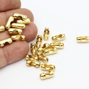May include: A close-up of a pile of gold-colored metal chain connectors. The connectors are small and cylindrical, with a twisted design. They are scattered on a white surface.