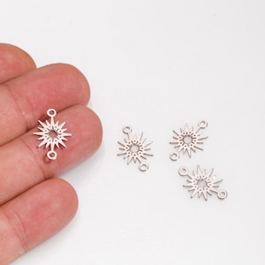 Puede incluir: Abalorios plateados con forma de estrella con dos bucles para hacer joyas. Los abalorios tienen un diseño calado detallado con rayos puntiagudos que emanan de un círculo central. Varios abalorios son visibles sobre un fondo blanco.