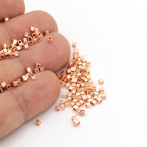 Könnte beinhalten: Nahaufnahme einer Hand, die einen Haufen kleiner, quadratischer, roségoldener Perlen hält. Die Perlen sind auf einer weißen Oberfläche verstreut.