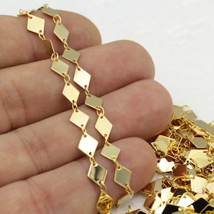 May include: Gold chain with square links. The chain is made of small, flat, square-shaped pieces of metal that are connected by small, round links. The chain is shown in a hand, with the ends of the chain hanging down.