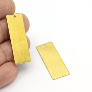 May include: Two rectangular brass metal tags with a small hole at the top. One tag is held by a hand. The tags are a light gold color and have a brushed finish. They are blank and ready for customization.