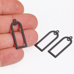 May include: Close-up of three black metal earring charms. The charms are open rectangles with a rounded top and a small loop for attaching to an earring hook. One charm is held in a hand.