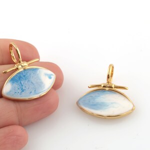 Puede incluir: Dos colgantes de tono dorado con un diseño de esmalte azul y blanco. Los colgantes tienen forma de ojo y tienen un bucle de tono dorado en la parte superior para sujetarlos a una cadena.