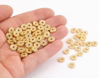 May include: A collection of small, gold-colored, textured beads with a circular shape and a hole in the center. Some beads are held in a hand, while others are scattered on a white surface. These beads are suitable for jewelry making.
