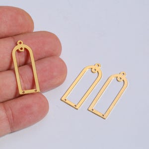Pode incluir: Pingentes de brincos de metal dourado em forma retangular arqueada. Os pingentes têm um topo decorativo e dois pequenos orifícios na parte inferior. Um pingente é segurado em uma mão e dois outros estão em um fundo claro.
