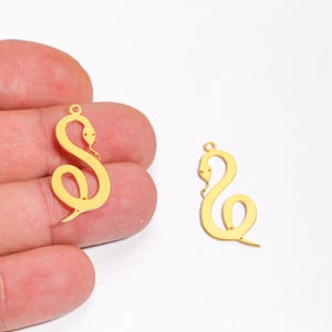 Puede incluir: Dos colgantes dorados con forma de serpiente. Los colgantes tienen un diseño estilizado, con un lazo en la parte superior para colgar. La cabeza de la serpiente es visible y el cuerpo está curvado. Los colgantes se muestran sobre un fondo blanco.