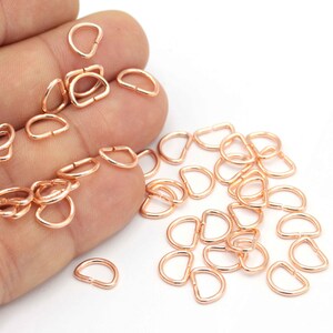 May include: A large pile of rose gold D-rings, also known as split rings, are scattered on a white surface. The rings are used for attaching charms, pendants, and other jewellery components.