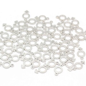 May include: A large quantity of silver metal charms in the shape of the male gender symbol. The charms are scattered on a white surface.