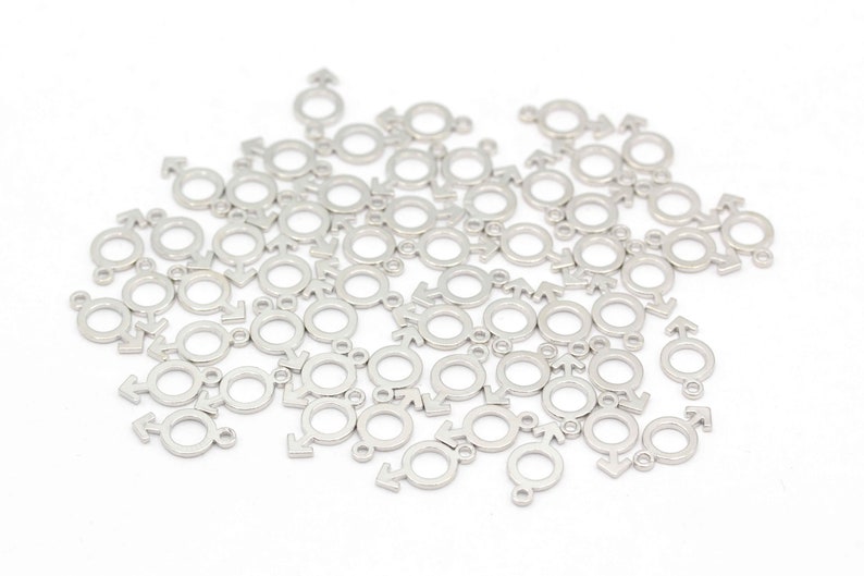 May include: A large quantity of silver metal charms in the shape of the male gender symbol. The charms are scattered on a white surface.