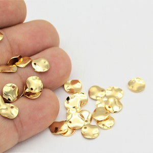May include: A group of gold-colored metal charms in the shape of a circle with a slightly irregular edge. The charms have two small holes at the top.