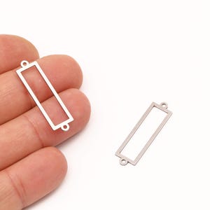 Op de afbeelding: Twee zilveren rechthoekige bedelhangers met kleine lussen aan elk uiteinde. Eén bedel wordt in een hand gehouden, de andere ligt op een wit oppervlak. Deze bedels zijn ideaal voor het maken van sieraden.