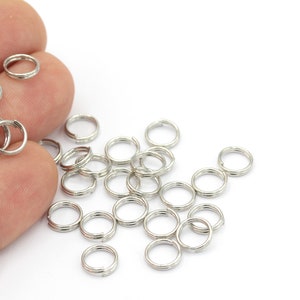 Peut inclure: Plusieurs petits anneaux fendus argentés, également appelés anneaux brisés ou anneaux de jonction, sont présentés sur un fond blanc. Ces anneaux à double boucle sont couramment utilisés en bijouterie et en artisanat.
