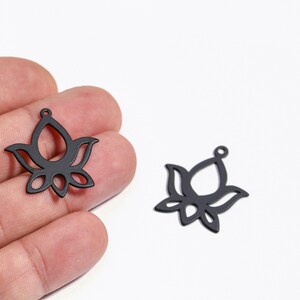 Può includere: Due ciondoli neri a forma di fiore di loto. Uno è tenuto in mano, l'altro è su una superficie bianca. I ciondoli hanno un design traforato con un piccolo anello in alto per il fissaggio. Ideale per la creazione di gioielli.