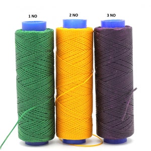 Peut inclure: Trois bobines de fil ciré, une verte, une jaune et une violette. Chaque bobine a un dessus en plastique bleu et le texte "1 NO", "2 NO" et "3 NO" imprimé sur le dessus.