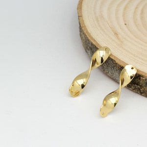Puede incluir: Dos piezas de metal retorcido de color dorado con un agujero en cada extremo. Las piezas están sobre una superficie de madera.