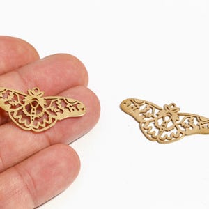 Puede incluir: Dos dijes de mariposa dorados, cortados con láser, con intrincados diseños calados. Uno se sostiene en una mano, mientras que el otro está sobre una superficie blanca. Los dijes presentan patrones de alas detallados y un pequeño lazo.