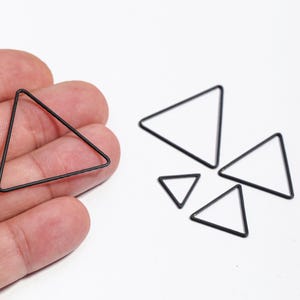 Puede incluir: Una colección de formas triangulares de metal negro de varios tamaños. Un triángulo se sostiene en una mano, mientras que otros están dispersos sobre una superficie blanca. Los triángulos tienen un diseño minimalista.