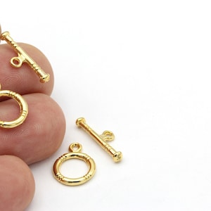 May include: Three gold-toned metal toggle clasps for jewelry making. Two clasps are round with a small loop at the top. The third clasp is a bar with two loops at one end.