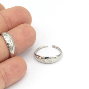 Puede incluir: Dos anillos de plata con un diseño decorativo. El anillo de la izquierda está en un dedo, mientras que el anillo de la derecha está sobre una superficie blanca.