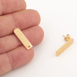 Könnte beinhalten: Zwei goldfarbene Ohrringrohlinge aus Metall mit gebürsteter Oberfläche. Jeder Rohling hat ein kleines Loch oben und einen Stift auf der Rückseite.