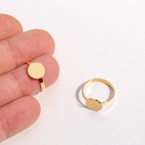 Puede incluir: Dos anillos ajustables dorados con una parte superior plana circular. Un anillo está en un dedo, el otro sobre una superficie blanca. Los anillos tienen un diseño simple y minimalista, adecuado para la fabricación de joyas o manualidades.