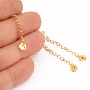 May include: Gold-colored necklace extenders with small, round charms. One extender has a charm with the letter "D", while the others have "E" and "H". The extenders are held in a hand against a white background.