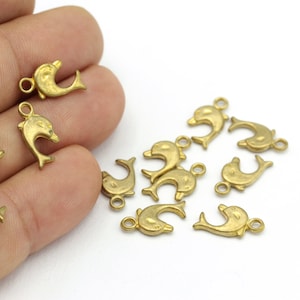May include: A group of gold-colored dolphin charms with jump rings. The charms are scattered on a white surface and a hand is holding some of the charms.