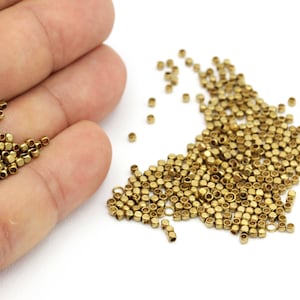 Puede incluir: Un montón grande de pequeñas cuentas de metal de color dorado. Las cuentas son hexagonales y tienen un agujero en el centro. Las cuentas están esparcidas sobre una superficie blanca.