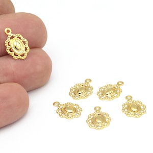Puede incluir: Encantos de colgante ovalados dorados con un borde calado decorativo. Los colgantes tienen un pequeño lazo en la parte superior para sujetarlos a las joyas. Varios colgantes están esparcidos sobre una superficie blanca.