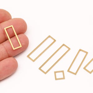Könnte beinhalten: Auf einer weißen Oberfläche sind mehrere goldfarbene, rechteckige und quadratische Metall-Schmuckzubehörteile zu sehen. Ein rechteckiges Zubehörteil wird von einer Hand gehalten. Die Zubehörteile sind unterschiedlich groß.