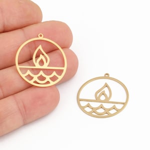 May include: Two gold-coloured metal pendants. Each pendant features a flame design within a circle, above a wavy line. The pendants have a small hole at the top for stringing. The pendants are on a white background.
