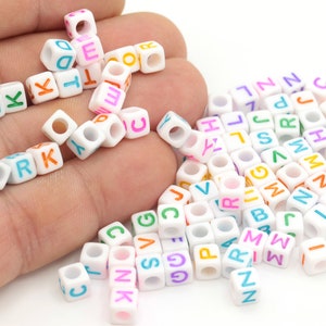 May include: A collection of white plastic letter beads with colorful letters. The beads are scattered on a white surface and a hand is holding some of the beads.