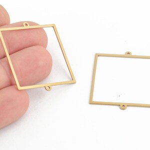May include: Two gold-coloured metal square frames with a loop at the top and bottom. The frames are empty and ready to be filled with resin or other materials.