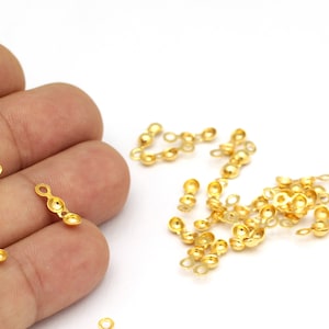 Könnte beinhalten: Nahaufnahme eines Haufens goldfarbener Metallperlenkappen mit einer Schlaufe an jedem Ende. Die Perlenkappen sind auf einer weißen Oberfläche verstreut. Einige der Perlenkappen werden von einer Hand gehalten.