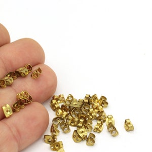 Peut inclure: Une collection de petits fermoirs d'oreilles dorés, également appelés papillons, est dispersée sur une surface blanche. Certains sont tenus dans une main, tandis que d'autres sont en vrac. Les fermoirs sont en métal et ont une forme de papillon.