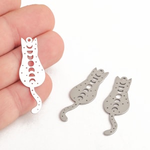 May include: Two silver metal cat charms with a moon phase design. The charms are in the shape of a cat with a long tail. The cat's body is filled with a pattern of crescent moons and stars.