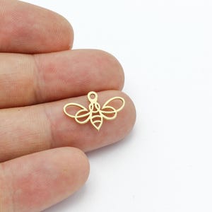 Könnte beinhalten: Goldfarbener Metall-Bienenanhänger mit einem Umrissdesign. Die Biene hat Flügel und einen Körper mit Streifen.