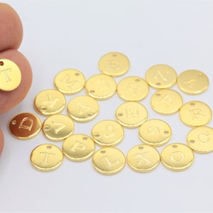 Puede incluir: Una colección de abalorios de letras del alfabeto de metal dorado. Cada abalorio es un círculo con una sola letra grabada en él. Los abalorios están esparcidos sobre una superficie blanca.