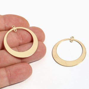 May include: Two gold-colored, open-circle earring components. Each component has a flat, crescent-shaped bottom and a small loop at the top for attachment. The earrings are shown against a neutral background.