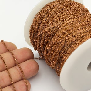 Könnte beinhalten: Nahaufnahme einer kupferfarbenen Kette mit kleinen, gleichmäßig verteilten Perlen. Die Kette wird auf einer weißen Spule präsentiert, wobei ein Teil der Kette herausragt. Die Kette wird wahrscheinlich für die Schmuckherstellung oder das Basteln verwendet.